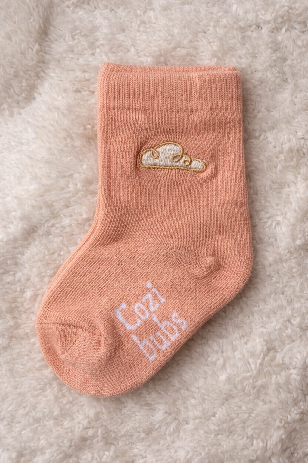Baby cotton sock in peach with Cozibubs logo and gold cloud embroidery on textured neutral background